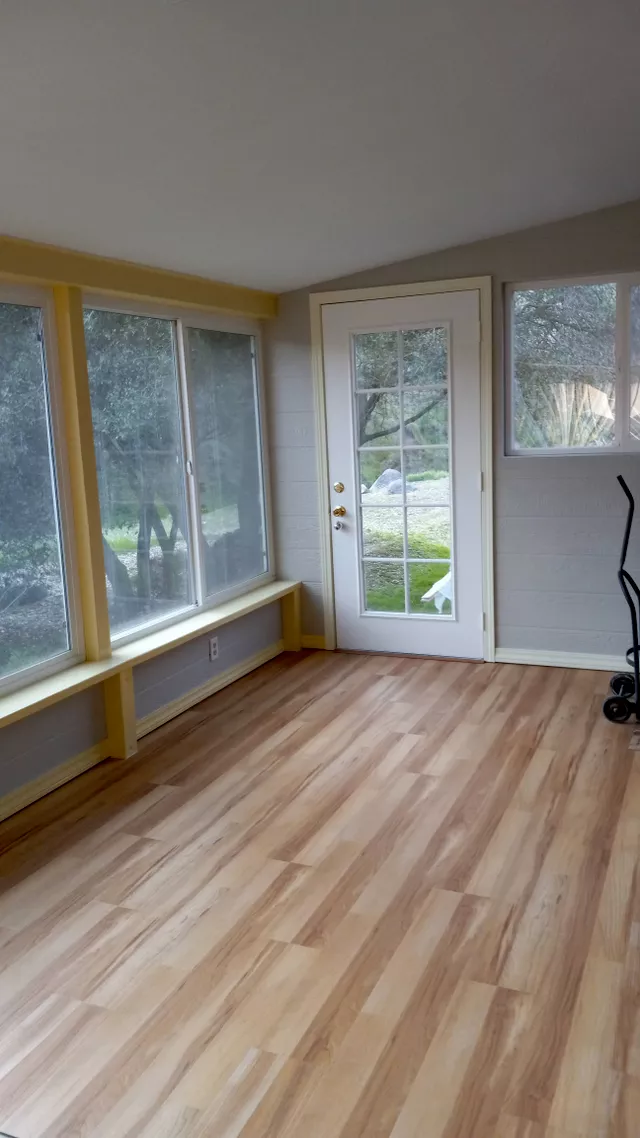 Bright sunroom with wood flooring, large windows, glass door, and yellow trim overlooking outdoor landscape